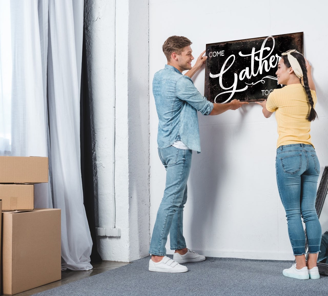 Modern Farmhouse Decor That newlyweds hanging up custom wall art in their new home. Moving boxes are in the background and couple is hanging a black sign with white lettering that says "gather". 