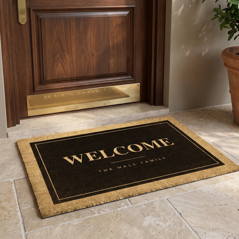 Black coir door mat reading Welcome with a family name, on a stone entry beside a terracotta pot