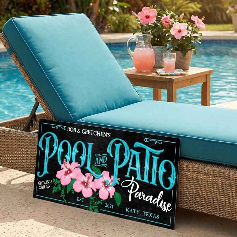 black pool decor sign leaning on a lounge chair with drink and flowers on the table beside a pool