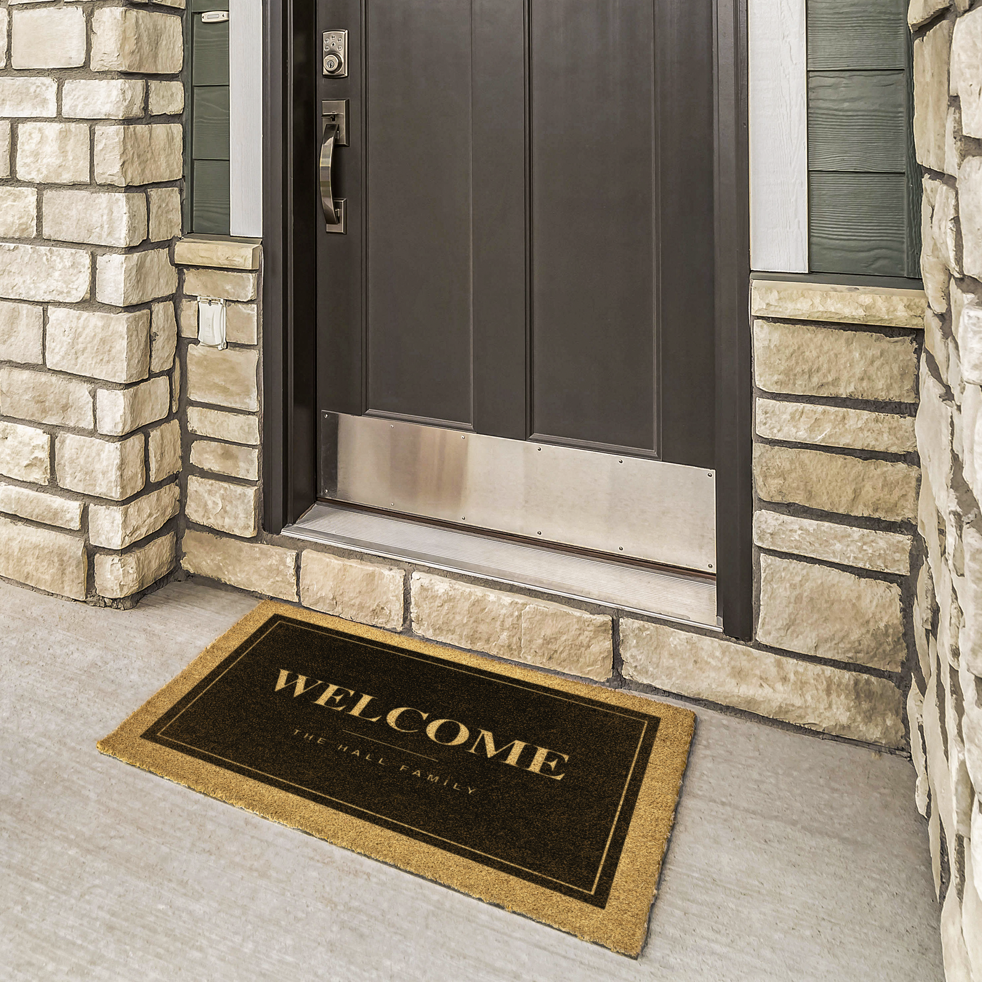 Black welcome mat with family name in cream at a bronze door framed by cream stone