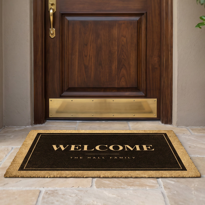 Black welcome mat with a family name in cream on a dark field, at a walnut door with a brass kickplate