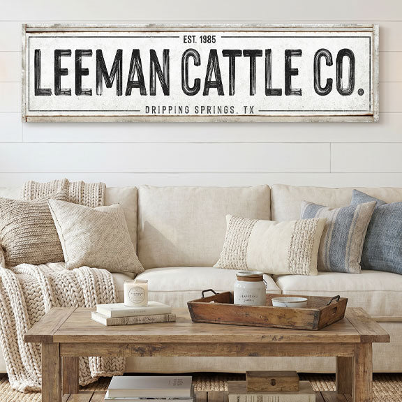 Living room with a beige sofa, wooden coffee table, and decorative pillows, featuring a large framed canvas cattle ranch sign above the sofa.