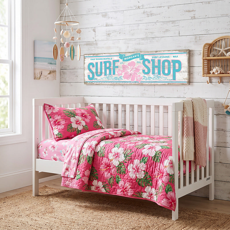 Coastal nursery decor, personalized surf shop sign above a white crib with pink hibiscus bedding and a shell mobile