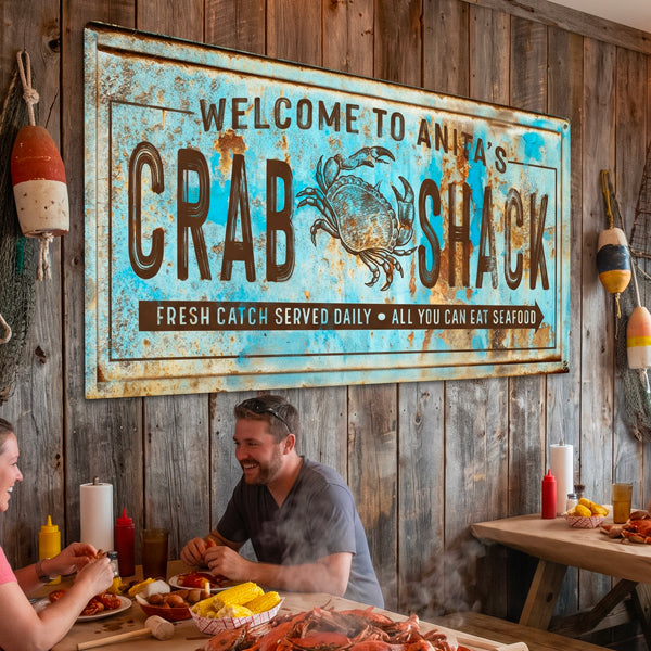 A large rustic metal wall sign reading 'Welcome to Anita's Crab Shack' with a crab illustration, mounted on a wood plank wall in a restaurant setting. Two people are seated at a table below, with seafood and condiments visible on the table and hanging buoys on the wall.