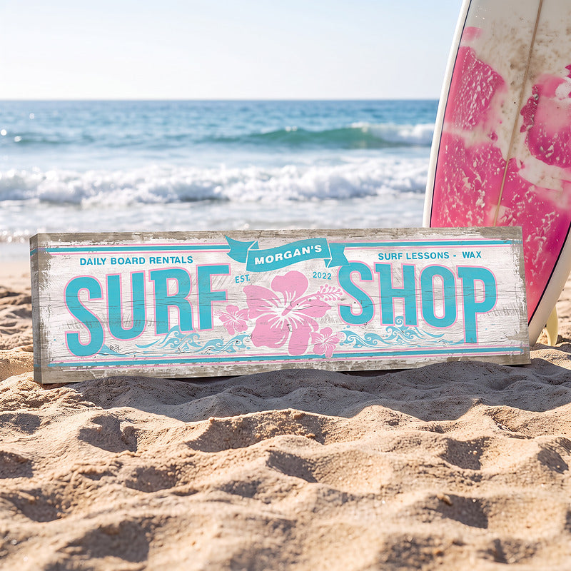 Coastal wall art surf shop sign standing in the sand by the ocean with a pink surfboard