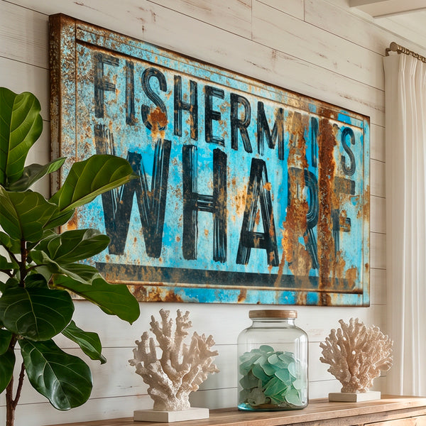 A rustic rectangular coastal wall decor sign with the words “FISHERMAN IS WHARF” in large black letters over a distressed blue and rust-colored background, displayed on a white shiplap wall above a shelf with coral and a glass jar.