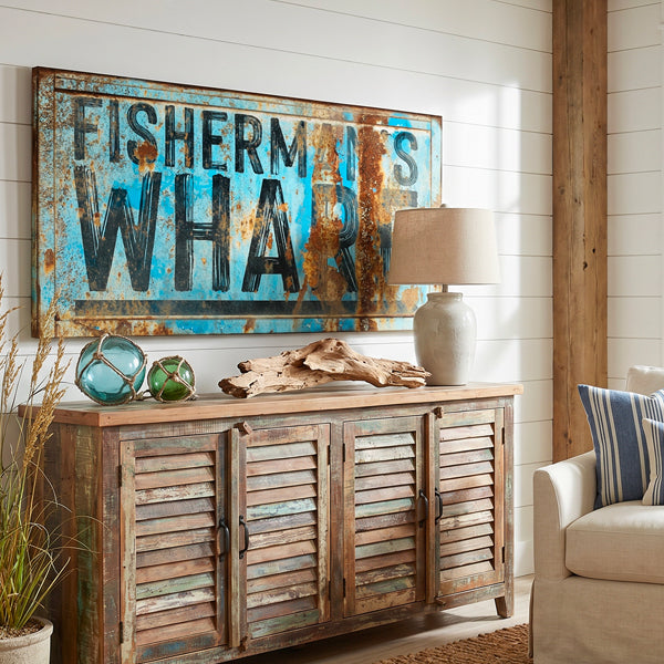 Coastal wall signs - Fisherman's Wharf' sign. The cabinet is topped with a table lamp, driftwood, and decorative glass spheres, with a striped armchair and potted plant visible nearby.