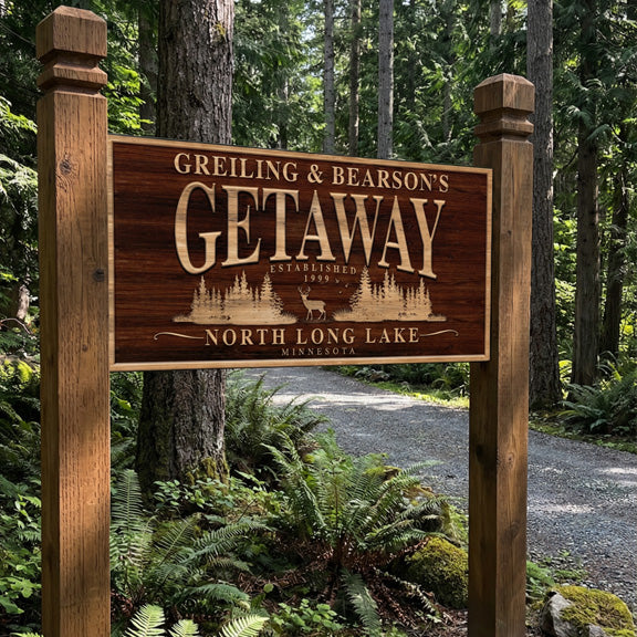Custom cabin sign mounted between cedar posts at a wooded driveway entrance with ferns