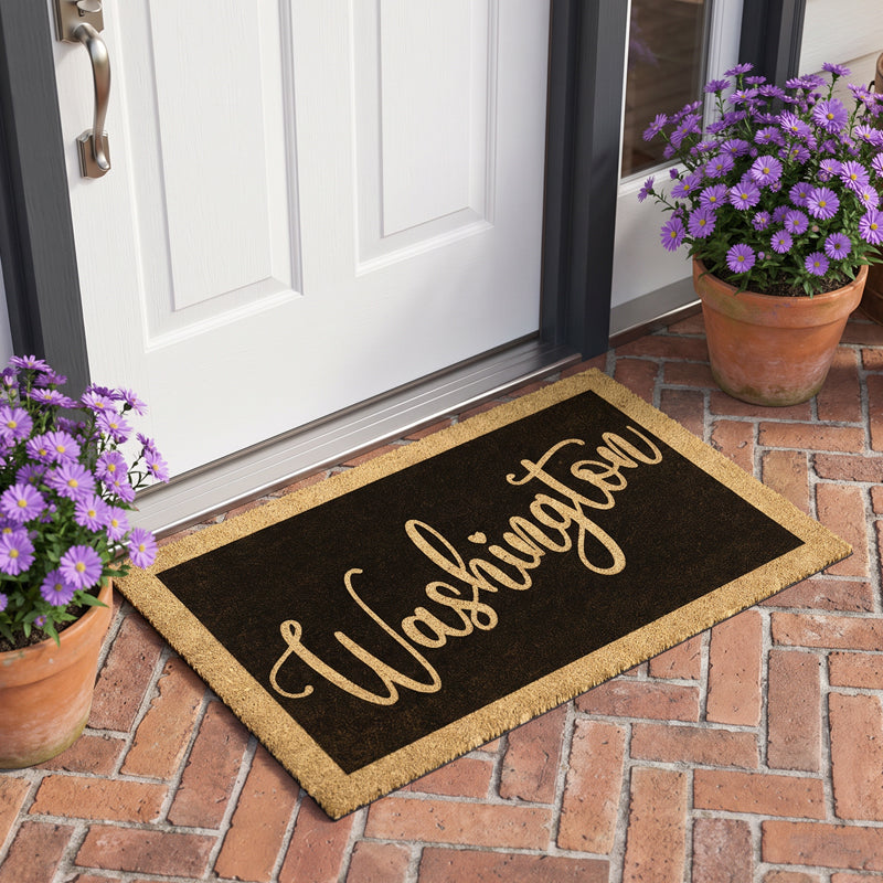 Custom surname door mat in gold script on black coir, on a brick porch between potted asters