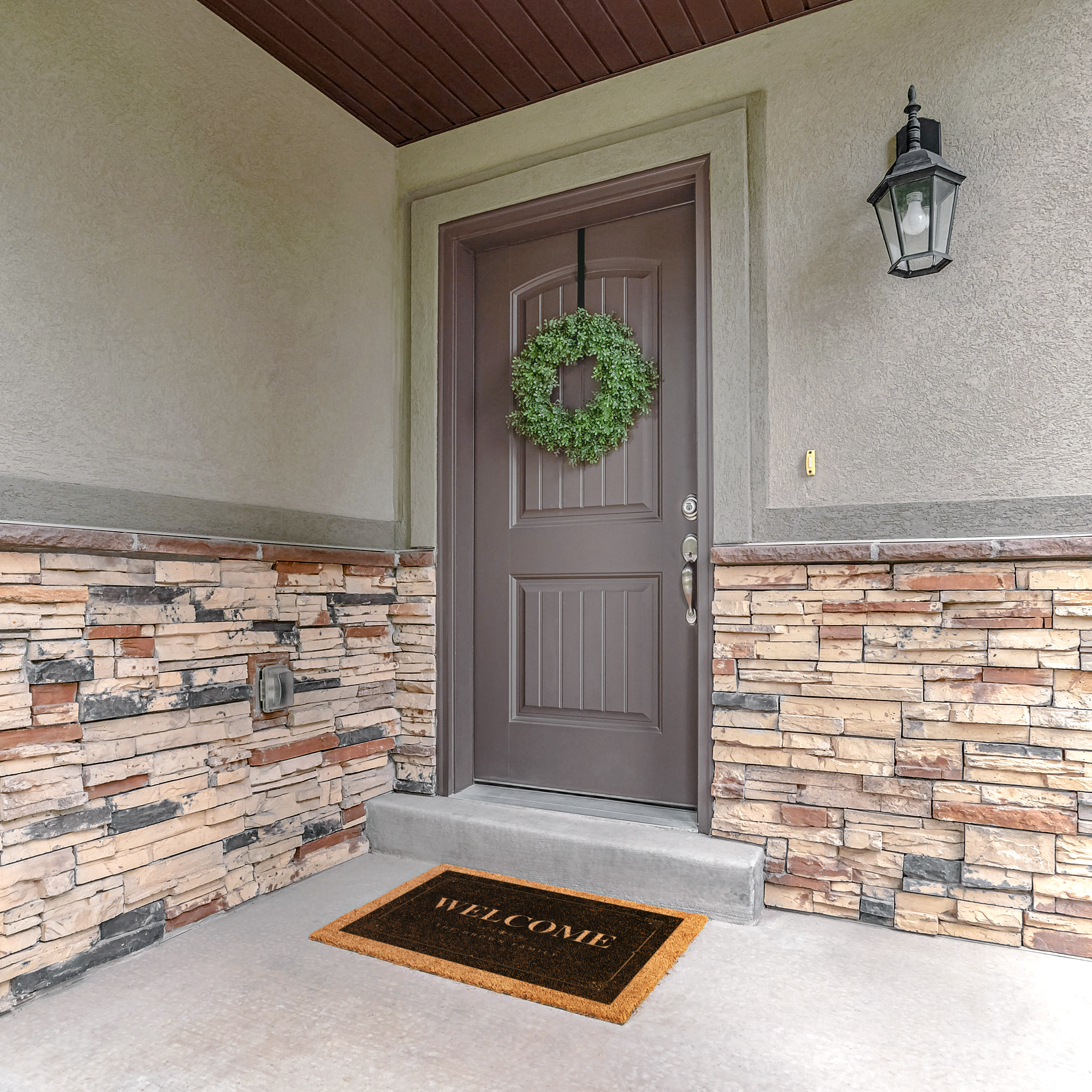Decorative door mat on a stone porch below a brown door with a green boxwood wreath