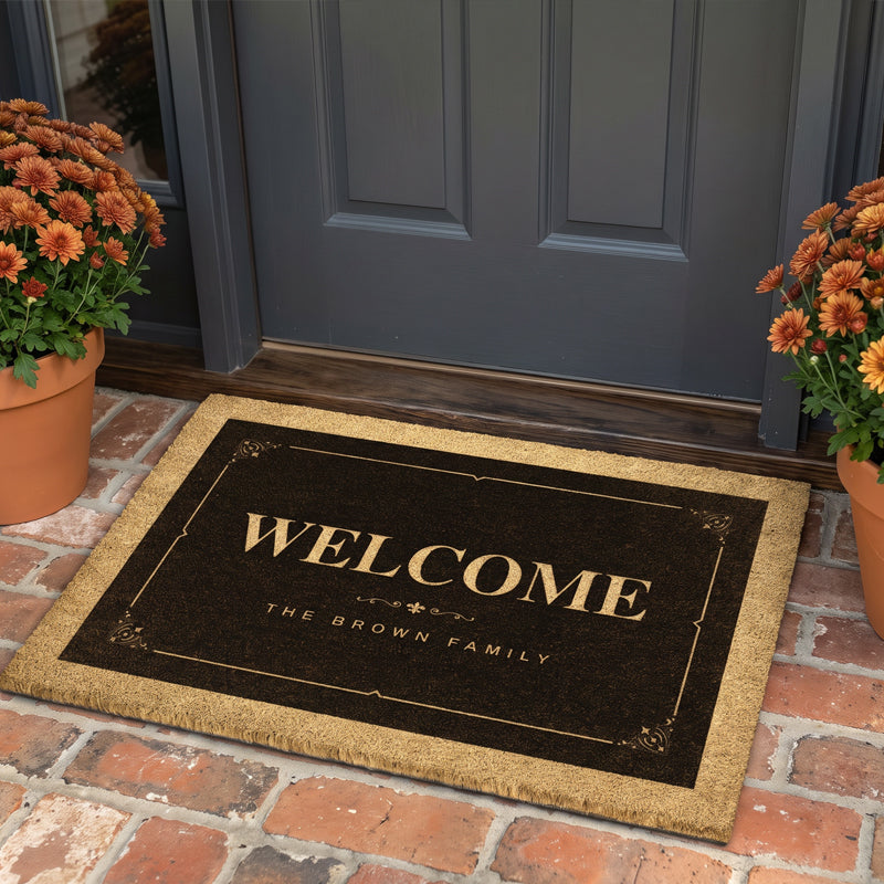 Decorative door mat reading Welcome with a family name in gold on brown, at a gray door with mums