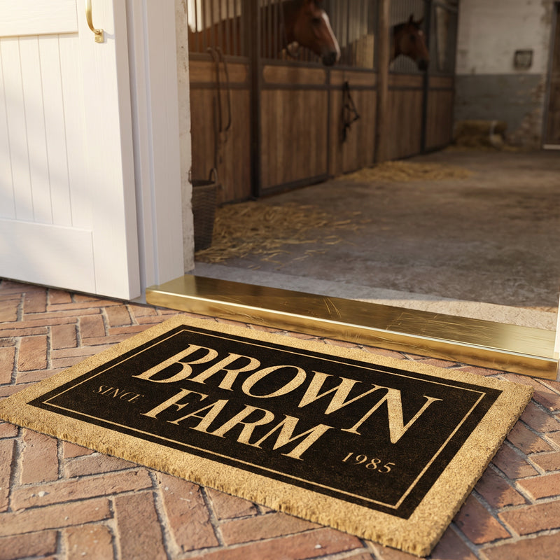 Equine doormat in cream on black coir at an open barn door, with two horses looking out of their stalls