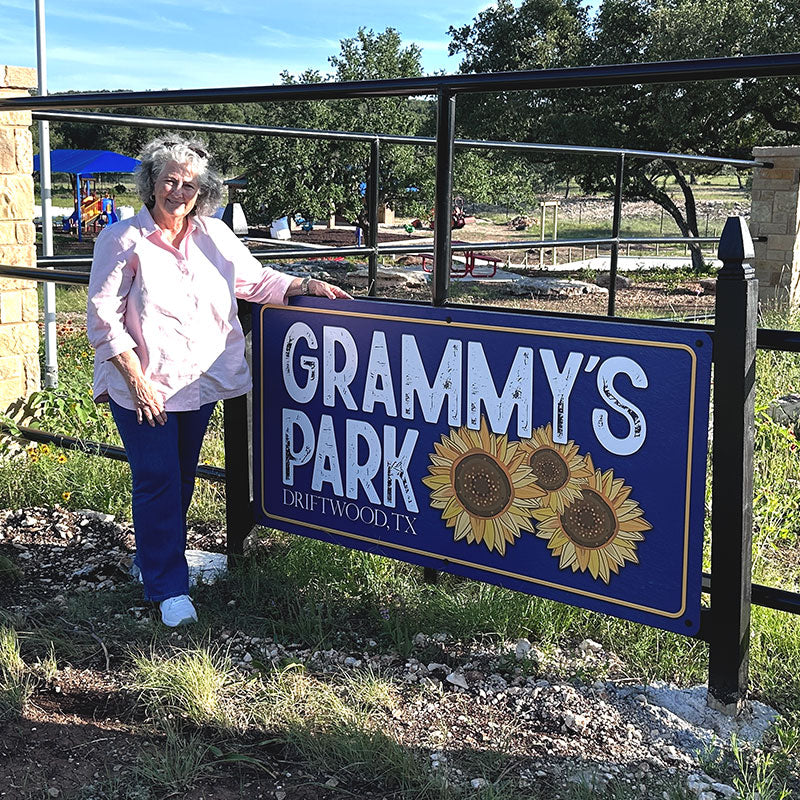 Grammy standing beside the blue and gold Grammy's Park entrance sign with sunflowers at the gate of a children's park in Driftwood, Texas