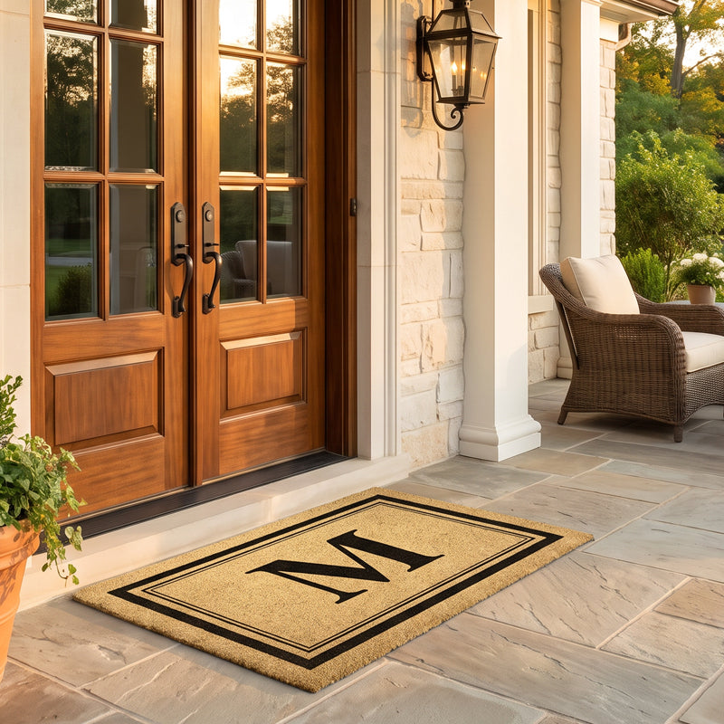 Initial doormat with monogram M on coir at double wood doors on a stone porch at sunset