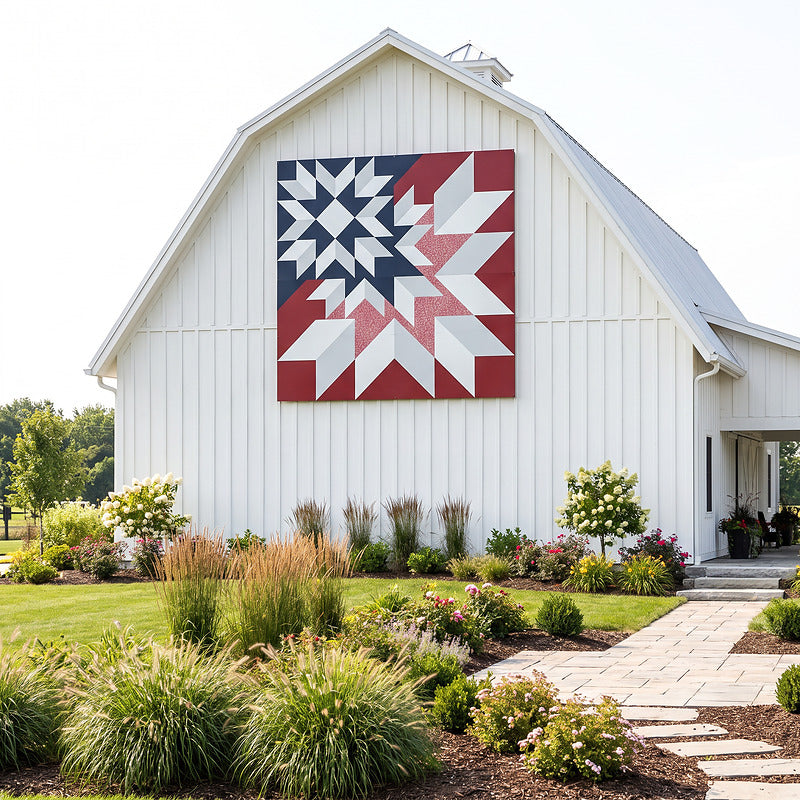 Metal barn quilt in red, white and blue mounted on the gable of a white gambrel barn above landscaped beds