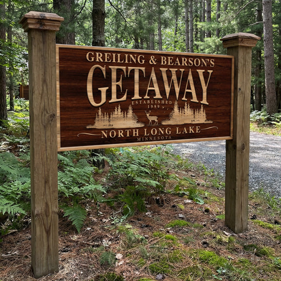 Metal lake house sign post-mounted between two posts at a wooded driveway entrance