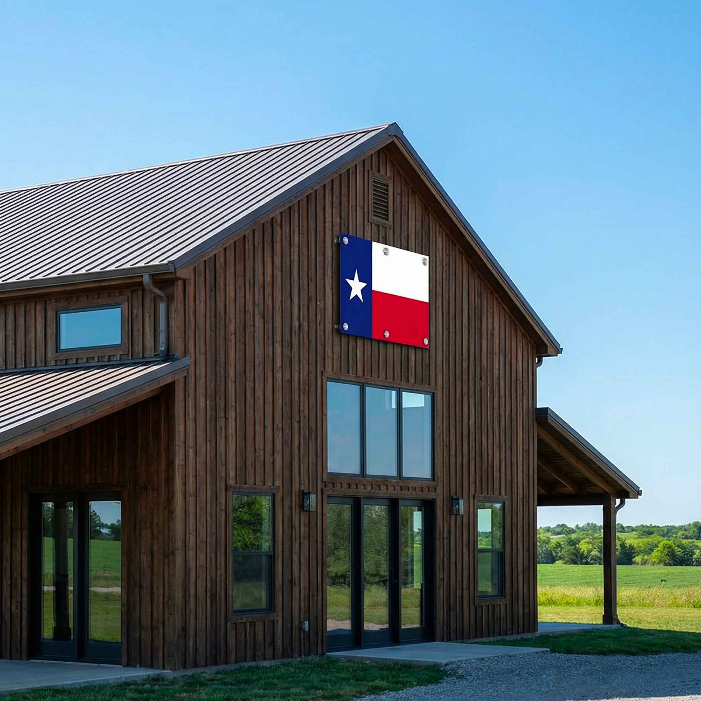 A large wooden barn-style building with a Texas flag mounted on the front gable, set against a clear blue sky with open fields visible on the right side.