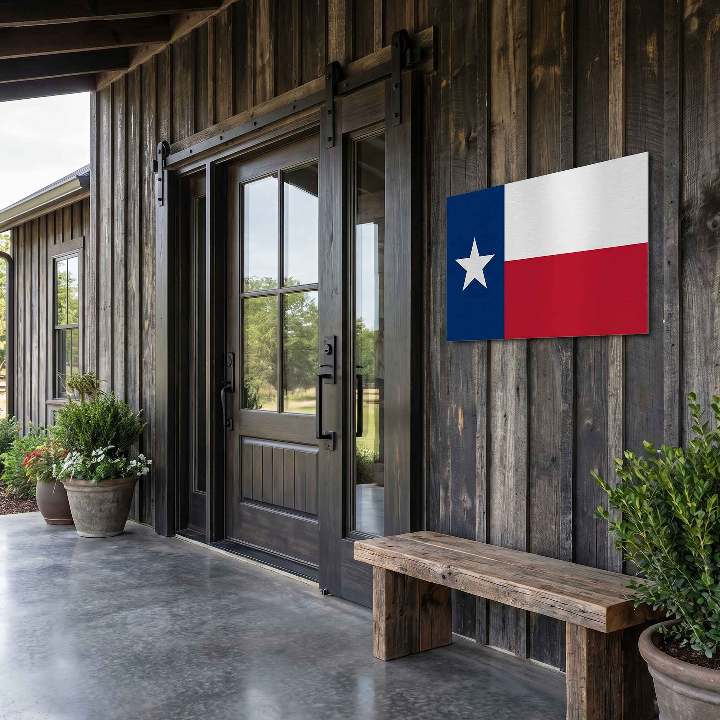Texas state flag wall art mounted on a dark wood plank exterior wall next to a wooden bench and potted plants on a porch.