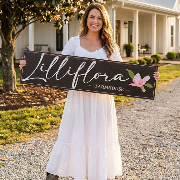 Woman holding a outdoor farm signs 'Lilliflora Farmhouse' sign in front of a house.