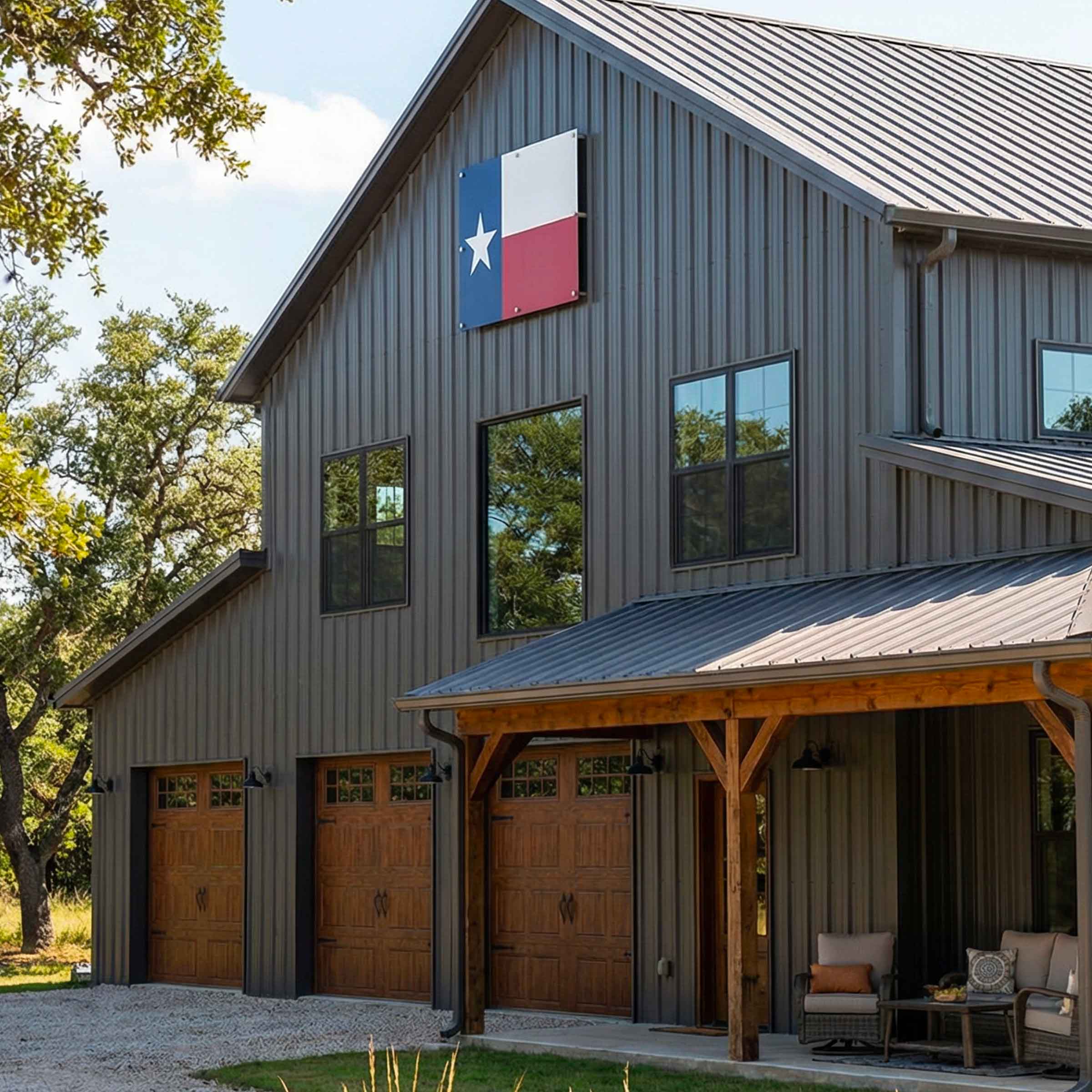 Outdoor metal texas flag displayed on a metal-sided barn-style building with three wooden garage doors, a covered porch with wooden posts, and a Texas flag mounted on the exterior wall. Trees and a gravel driveway are visible around the structure.