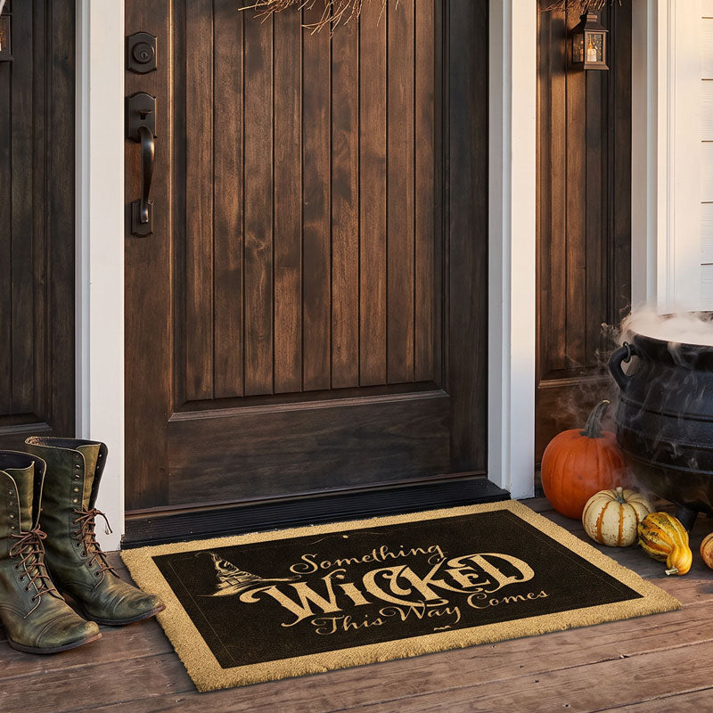 Something Wicked This Way Comes doormat at a dark wood door with pumpkins and a steaming cauldron