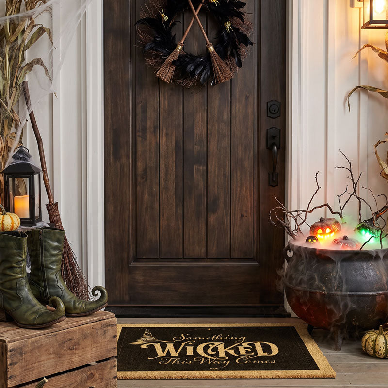 Something Wicked welcome mat below a black broom wreath with a glowing cauldron and cornstalks
