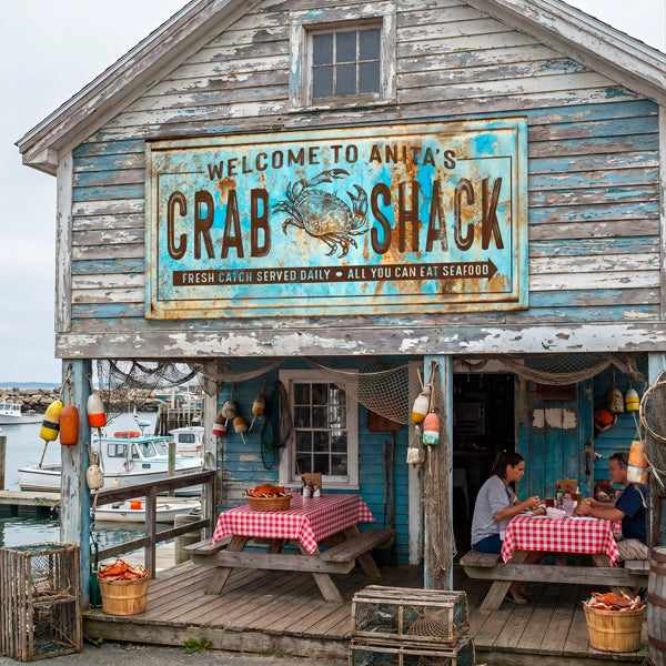 vintage coastal decor mounted on a  building labeled 'Welcome to Anita's Crab Shack' with a large weathered sign featuring a crab illustration. The front porch has picnic tables with red-and-white checkered tablecloths, hanging fishing nets and buoys, and two people seated under the porch.