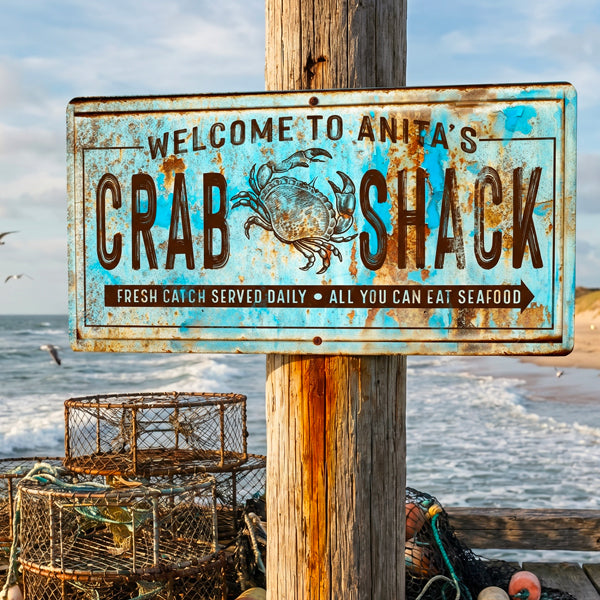 Vintage Crabsign reading “Welcome to Anita’s Crab Shack” with a blue distressed background, brown lettering, and a crab illustration, mounted on a wooden post at the beach with lobster traps and ocean waves behind it.