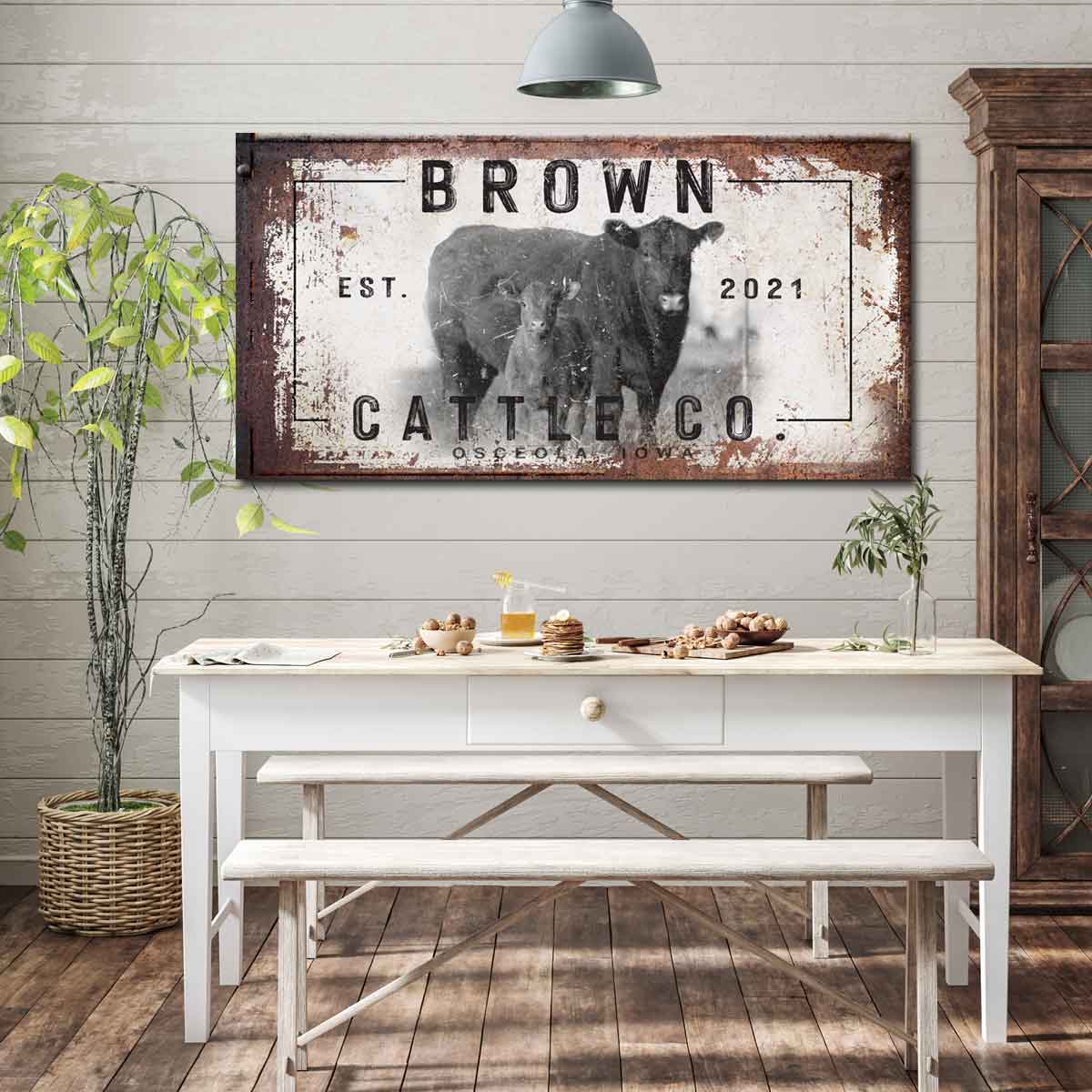 Cattle Ranch canvas sign with Red Angus cows standing in front. Words Brown Cattle Company, with establish date on distressed rusty frame and white peeled paint cow print.