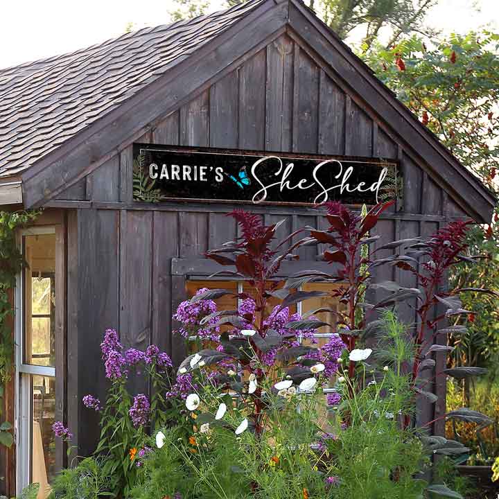 Metal She shed sign on rustic black background with white script lettering and a butterfly.